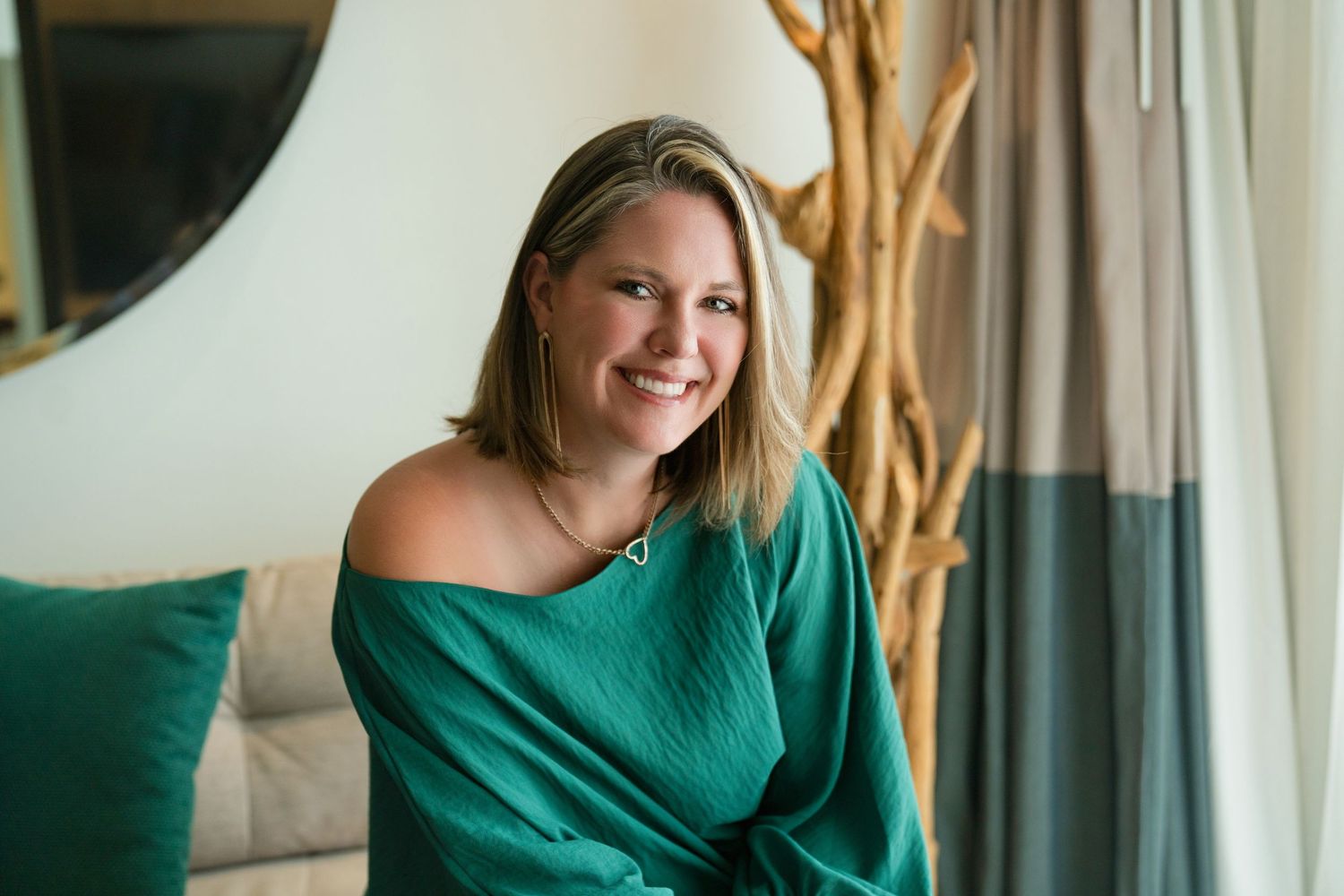 Woman in emerald green dress poses elegantly in front of window with natural lighting.