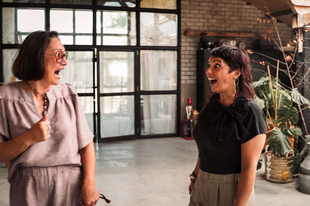 A group of people laughing and socializing in a bright industrial-style space with brick walls and large windows.
