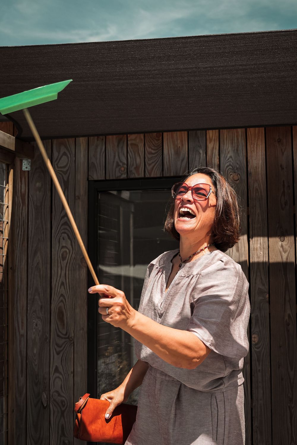 A joyful figure in a gray dress holds a green broom while laughing against a dark wooden wall.
