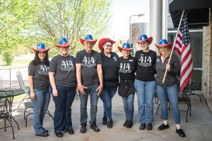 A line of people wearing matching blue shirts and jeans stand together under an outdoor covered area with American flags.