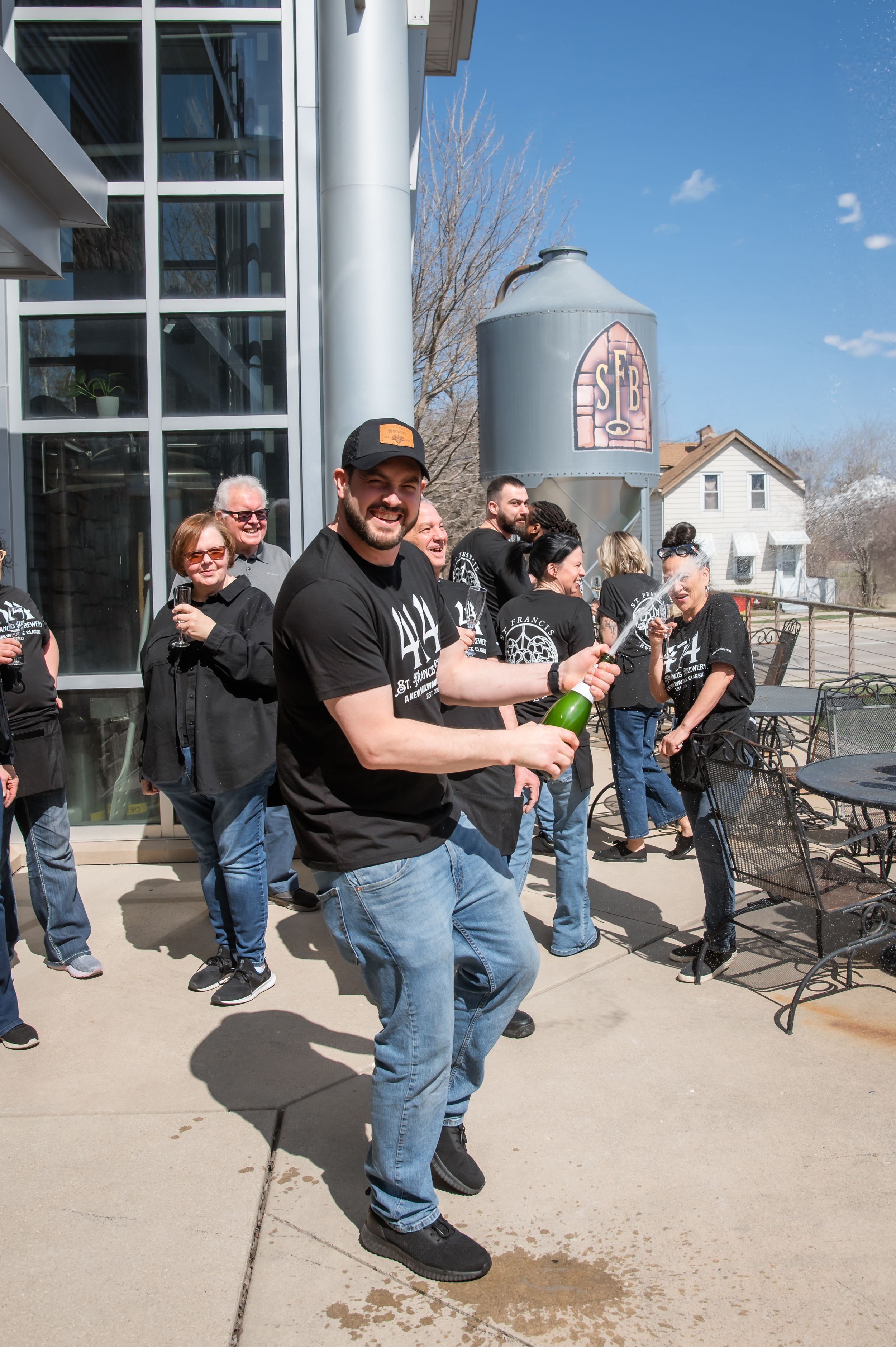 A group gathers outside a brewery with a large metal silo tank visible in the background on a sunny day.
