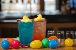 Colorful cocktails garnished with rubber ducks and balloons sit on a dark wooden bar counter.