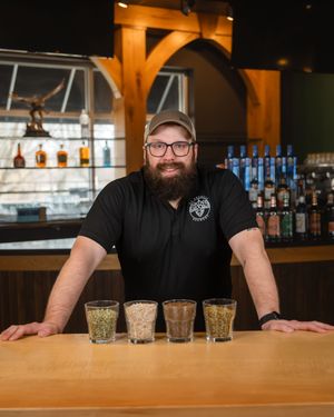 A bartender stands behind a wooden bar counter with four empty glasses lined up in front.