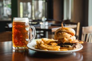 A juicy hamburger with french fries served on a white plate alongside a tall glass of amber beer at a restaurant table.