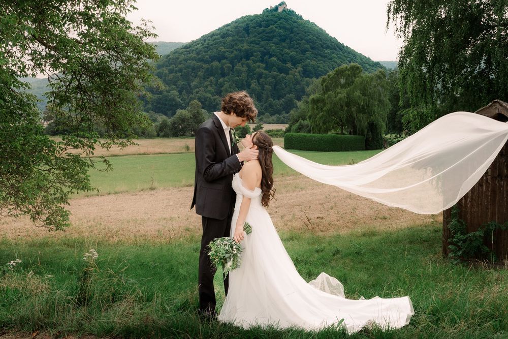 Wedding couple posing in scenic mountain landscape with flowing veil and rustic barn.