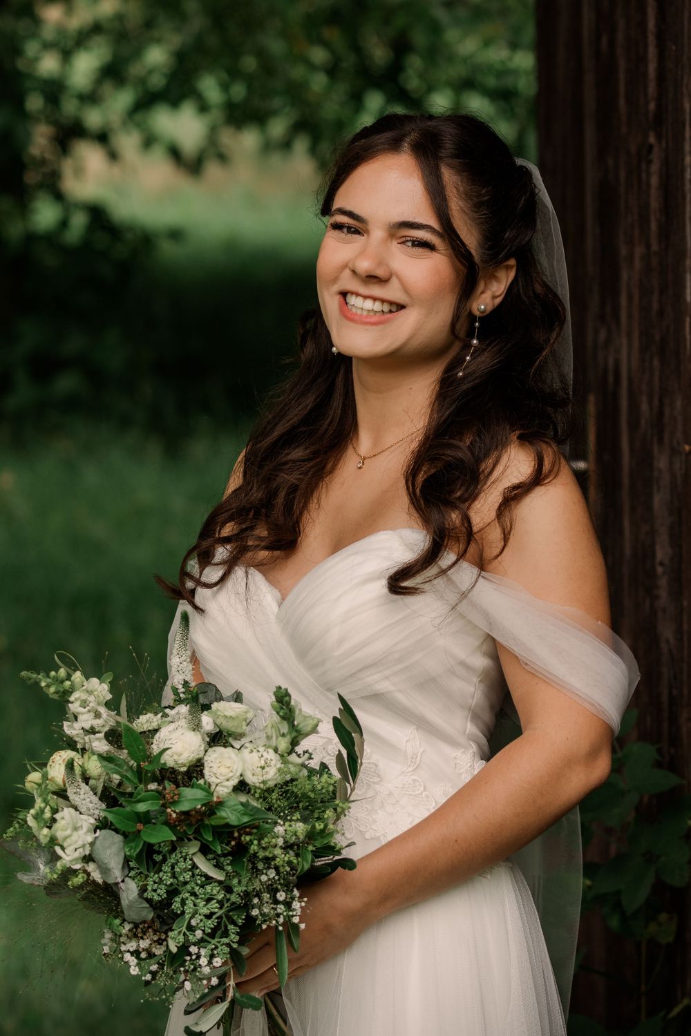 A beautiful bride in a white off-shoulder dress holds a green and white bouquet against a natural outdoor backdrop.