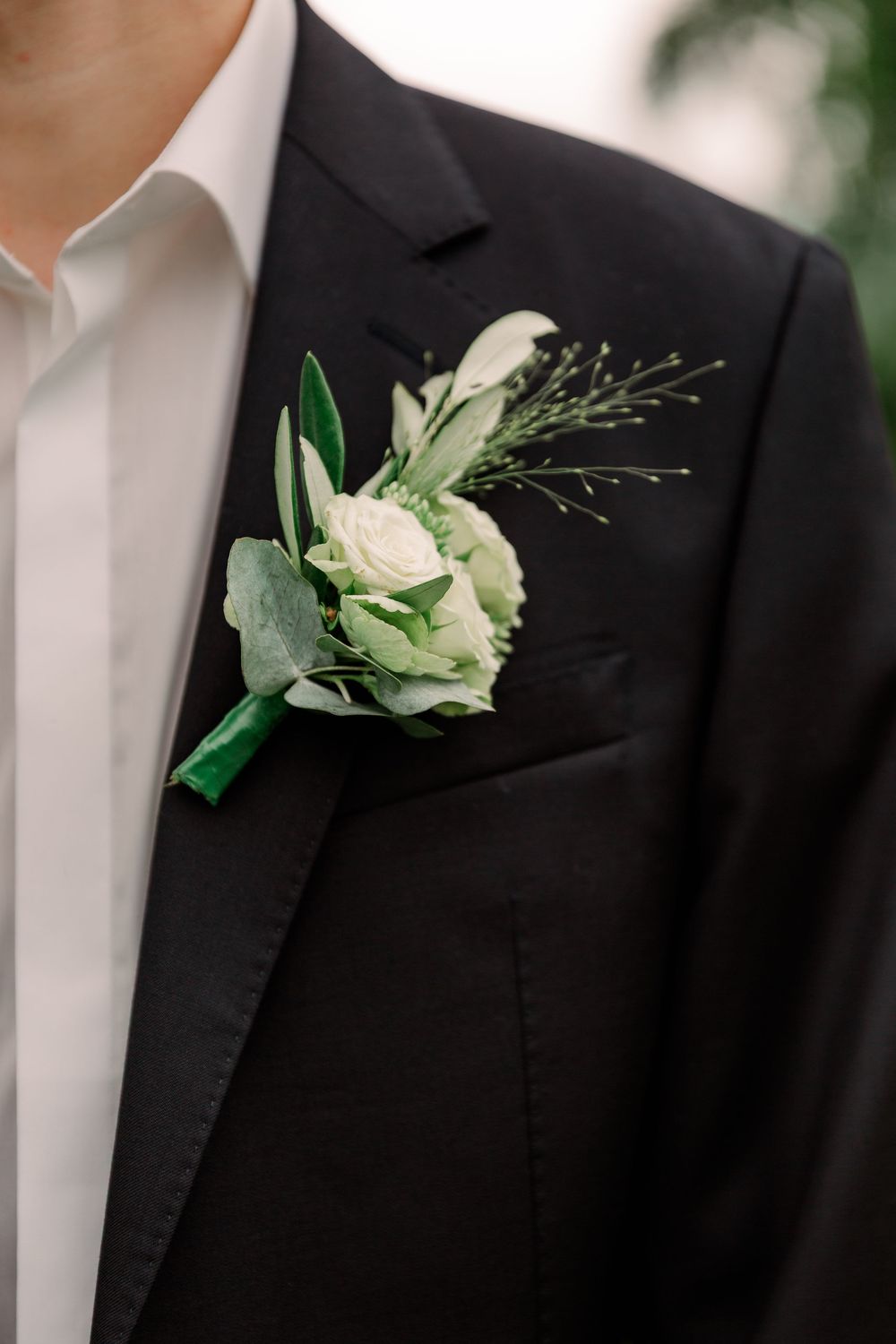 White rose boutonniere with greenery pinned to a black suit lapel.
