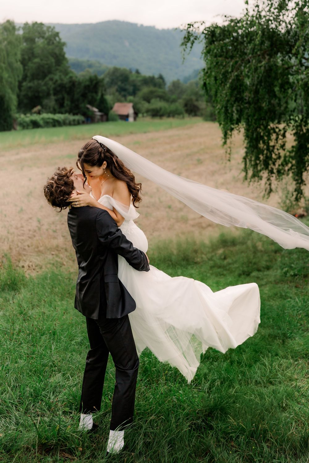 A joyful wedding couple dancing in a green field with mountains in the background.
