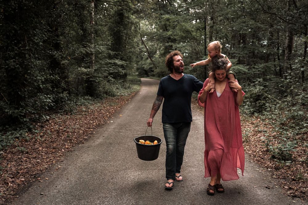 Two people walk along a forested path carrying a black bucket on an autumn day.