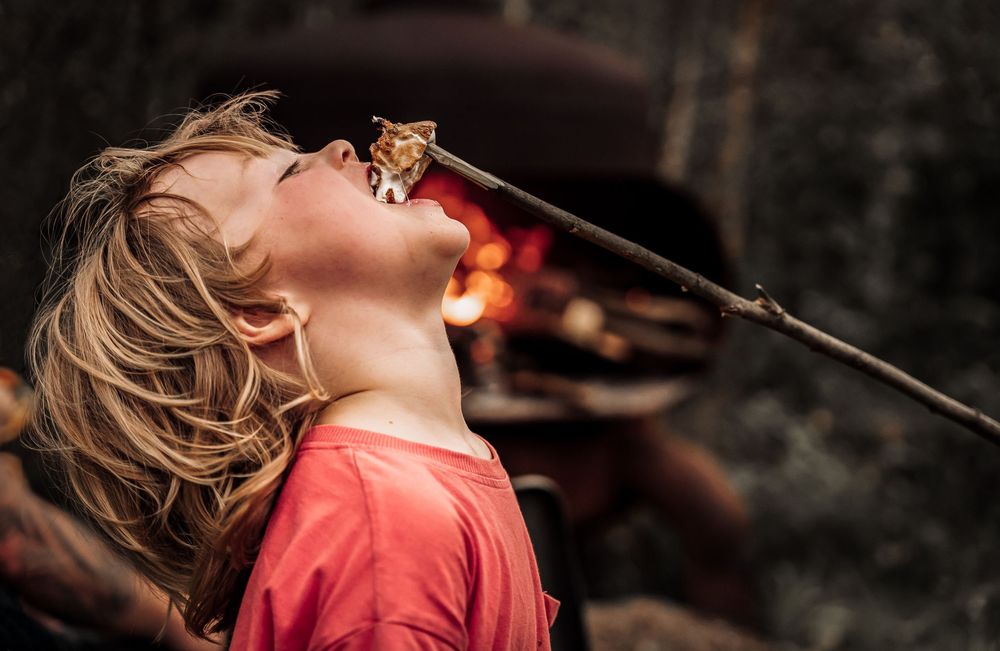 Child in red shirt with head tilted back near glowing marshmallow roasting stick at dusk.