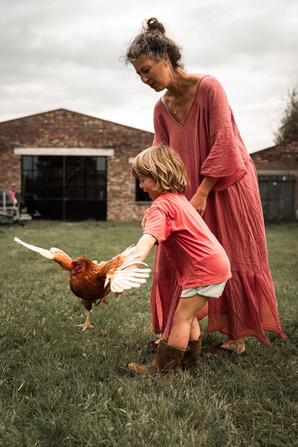A rustic farm scene shows feeding a rooster in a pink dress and boots near a brick building.