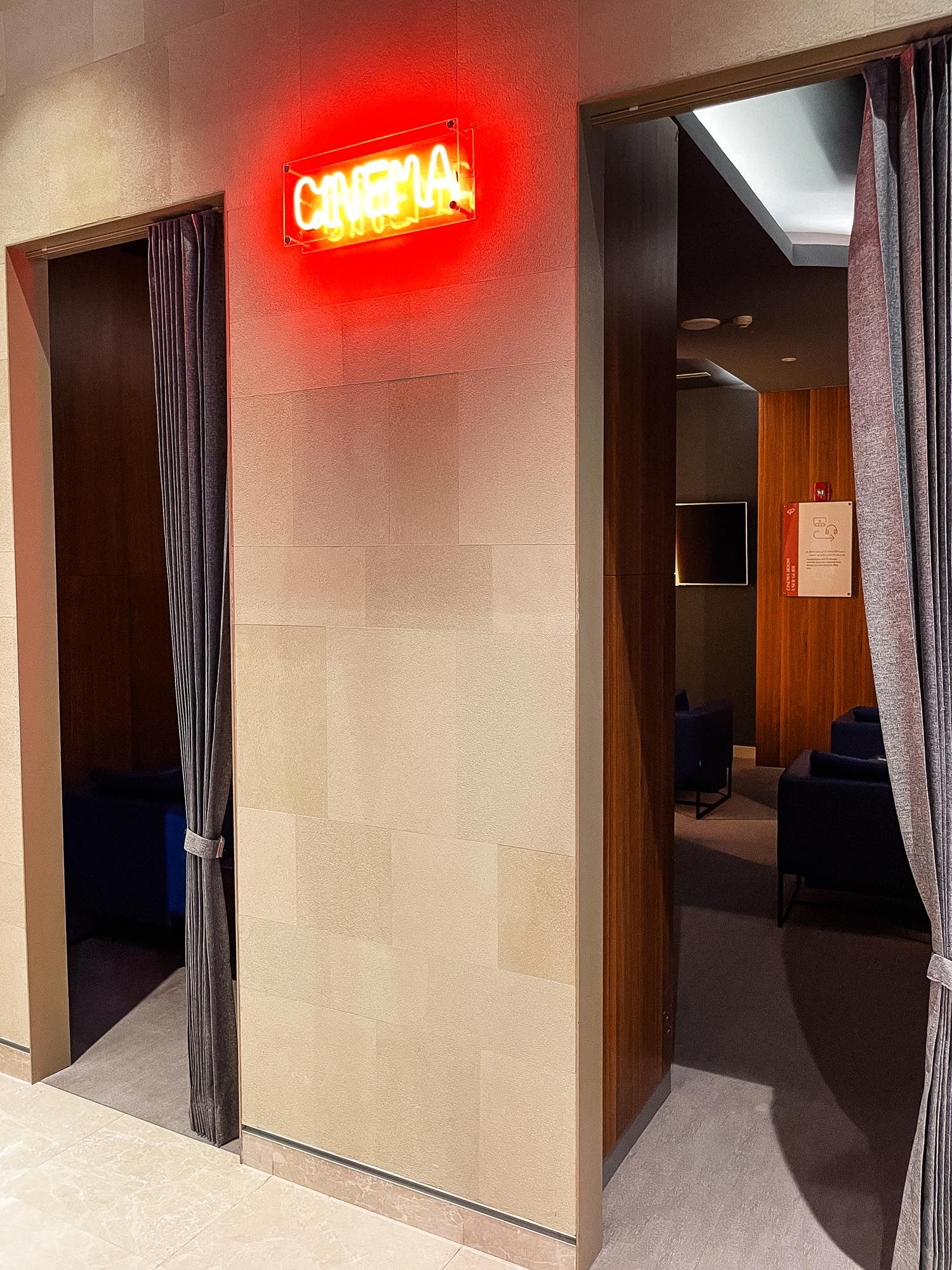 Dimly lit hotel hallway with beige walls, dark curtains, and a glowing red neon sign at the end of the corridor.