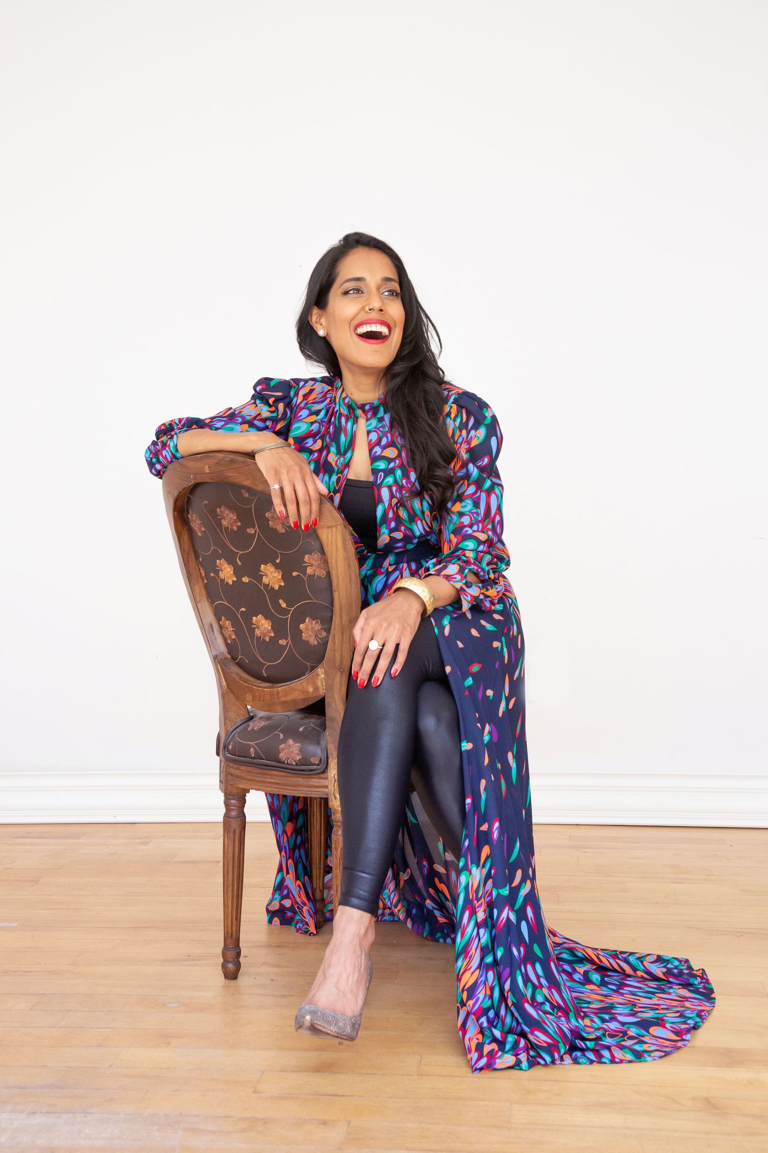 Professional woman in green blazer and white graphic t-shirt sits confidently on white chair against white background.