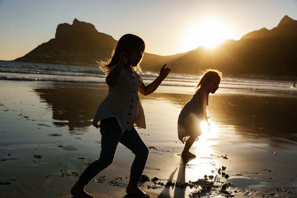 Silhouetted figures play and explore on a scenic beach at sunset with golden light reflecting off the sand and water.