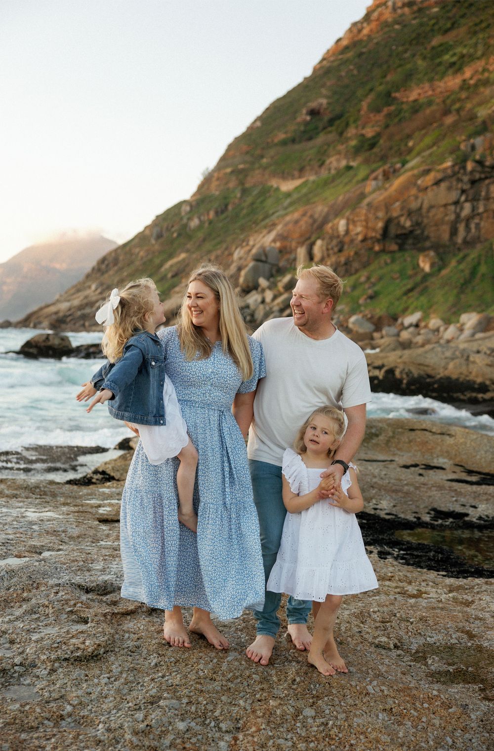 Family in coordinating blue and white outfits walking together on a rocky beach at sunset with coastal cliffs in background.
