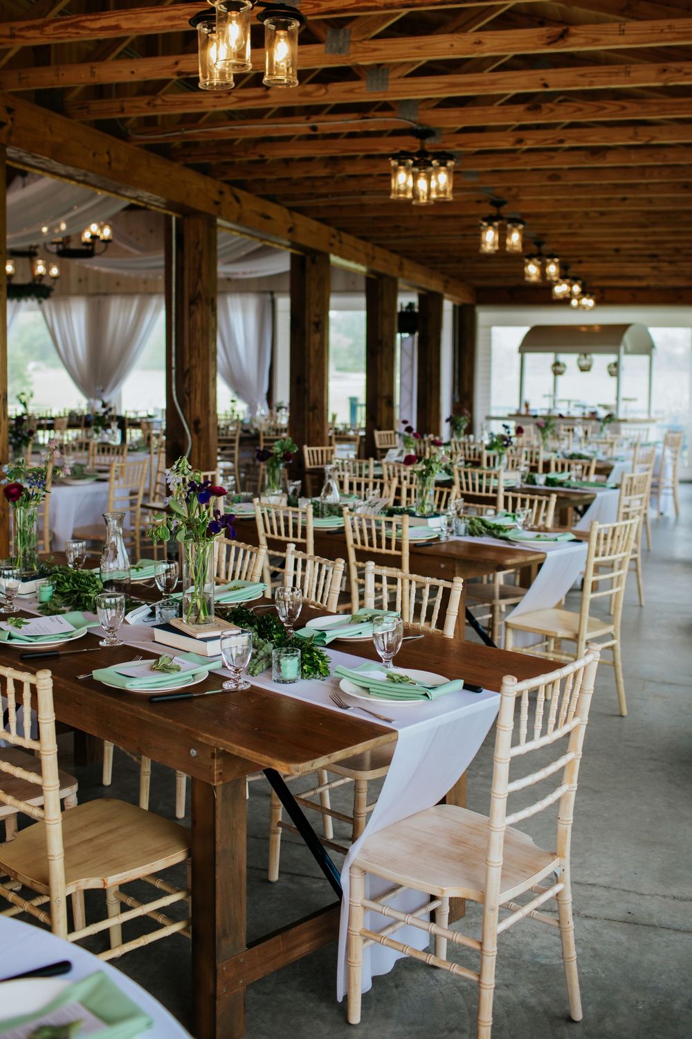Rustic wedding reception venue with wooden farm tables, white chiavari chairs, and hanging pendant lights.