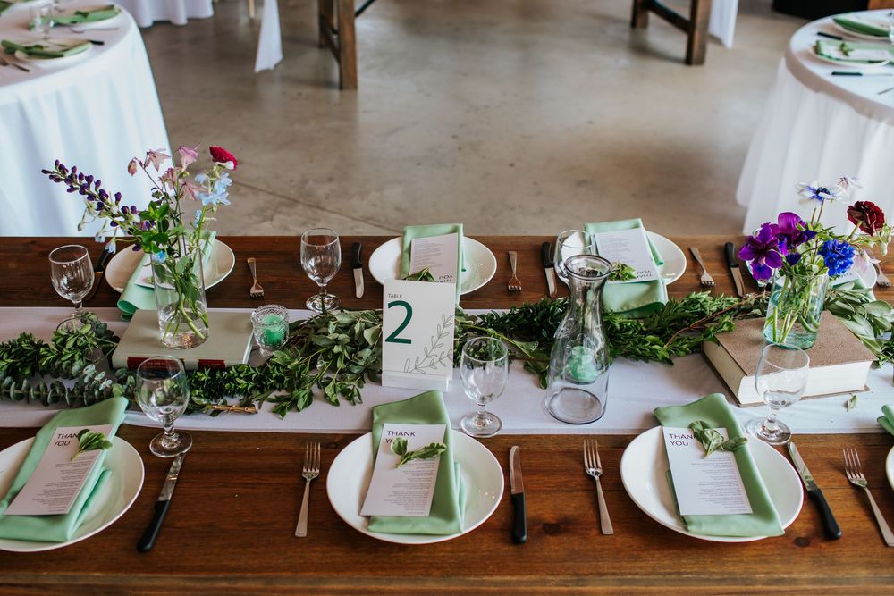 Elegant table settings with greenery and purple floral accents at rustic wedding reception.