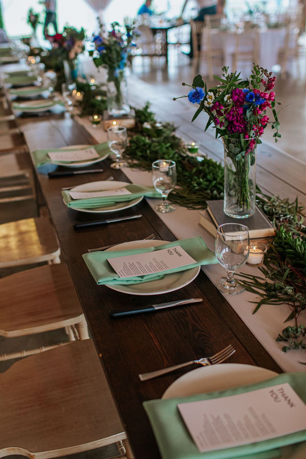 Elegant wedding reception table setting with green accents, white plates and floral centerpieces on dark wood tables.