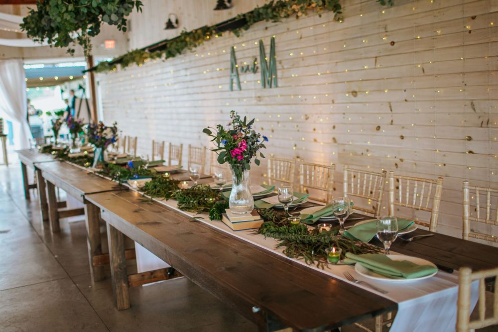 Long wooden farm tables arranged in a rustic barn venue with white brick walls and hanging greenery.