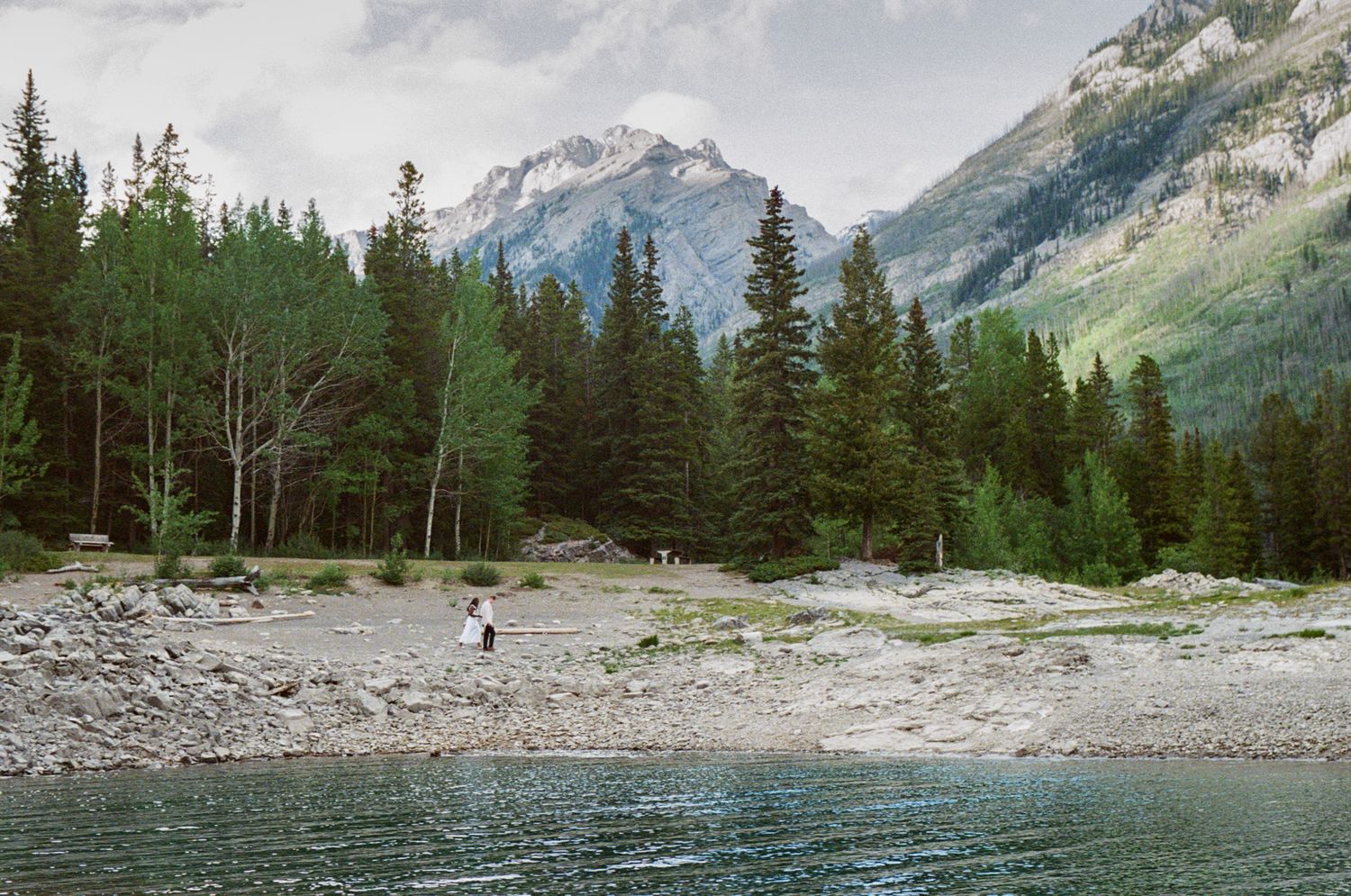 35 mm film photo of bride and groom wandering the shoreline of Lake Minnewanka in Banff with mountains in the background