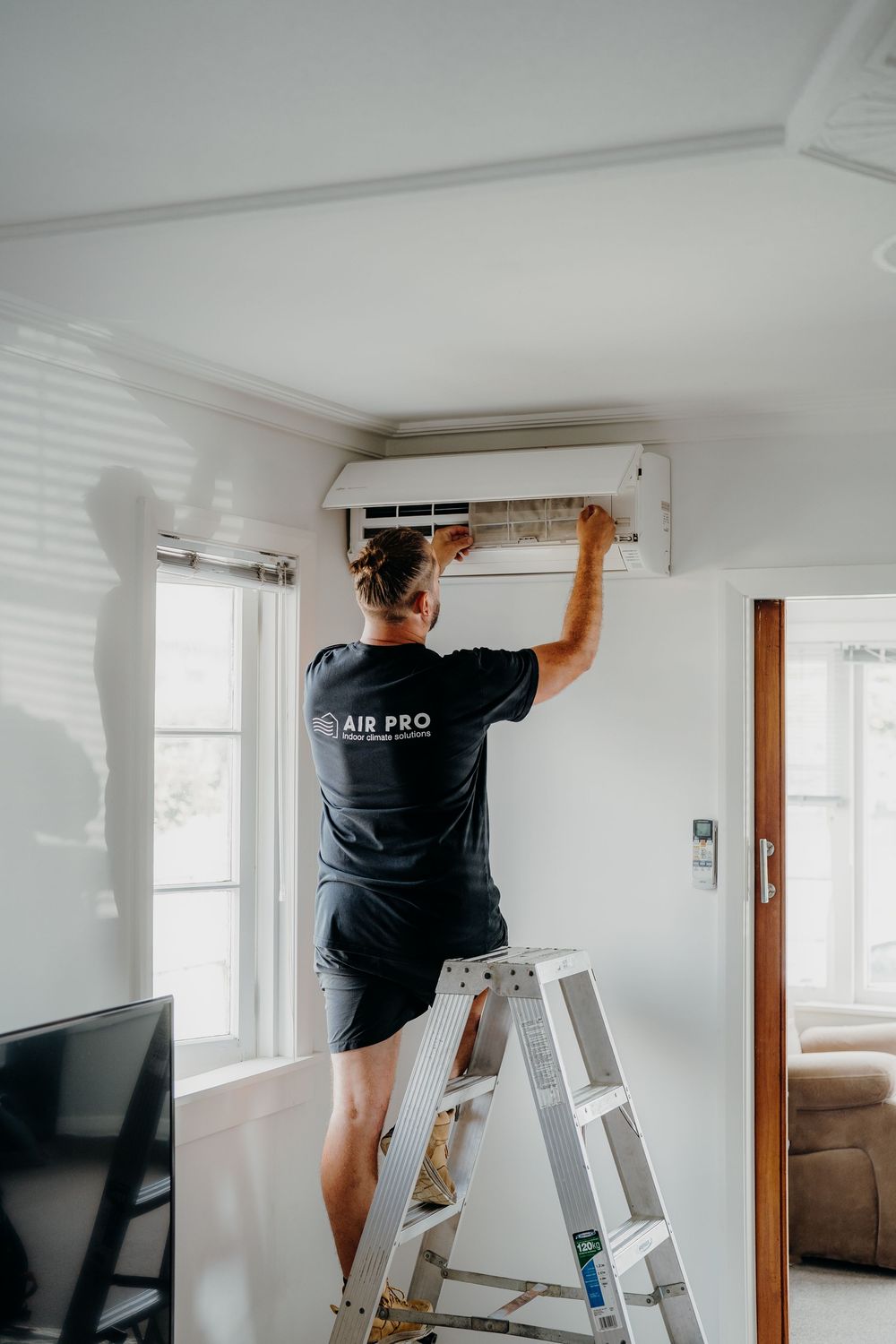 HVAC technician on ladder servicing an indoor air conditioning unit mounted on white wall.