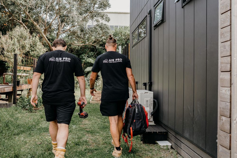 Two air conditioning technicians in black uniforms walking on grass path outside residential building.
