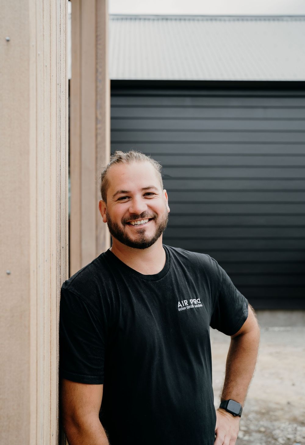 A person wearing a black t-shirt smiles warmly against a light-colored background in a casual portrait series.