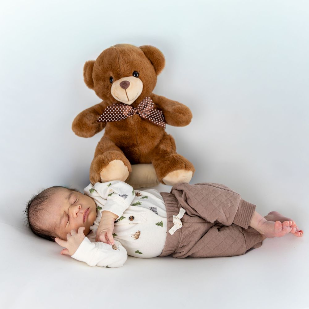 Sleeping baby in white and brown outfit lying next to brown teddy bear with checkered bow on white background.