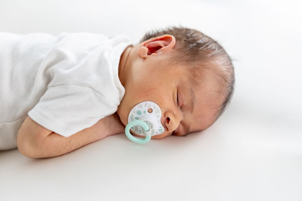 A sleeping baby in white clothing with a pacifier lies peacefully on a white surface.