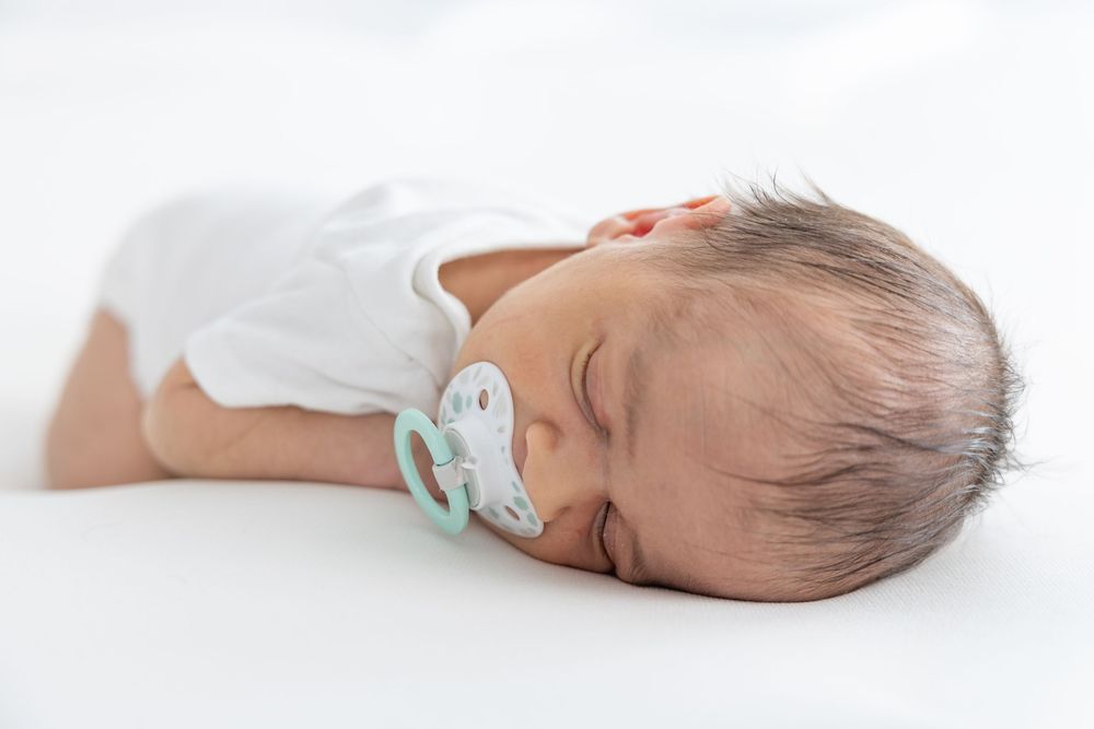 Peaceful sleeping baby wearing white onesie with mint green pacifier on white background.