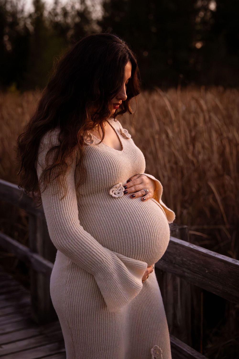 A pregnant figure in a white knit dress stands on a wooden boardwalk at sunset among tall grasses.