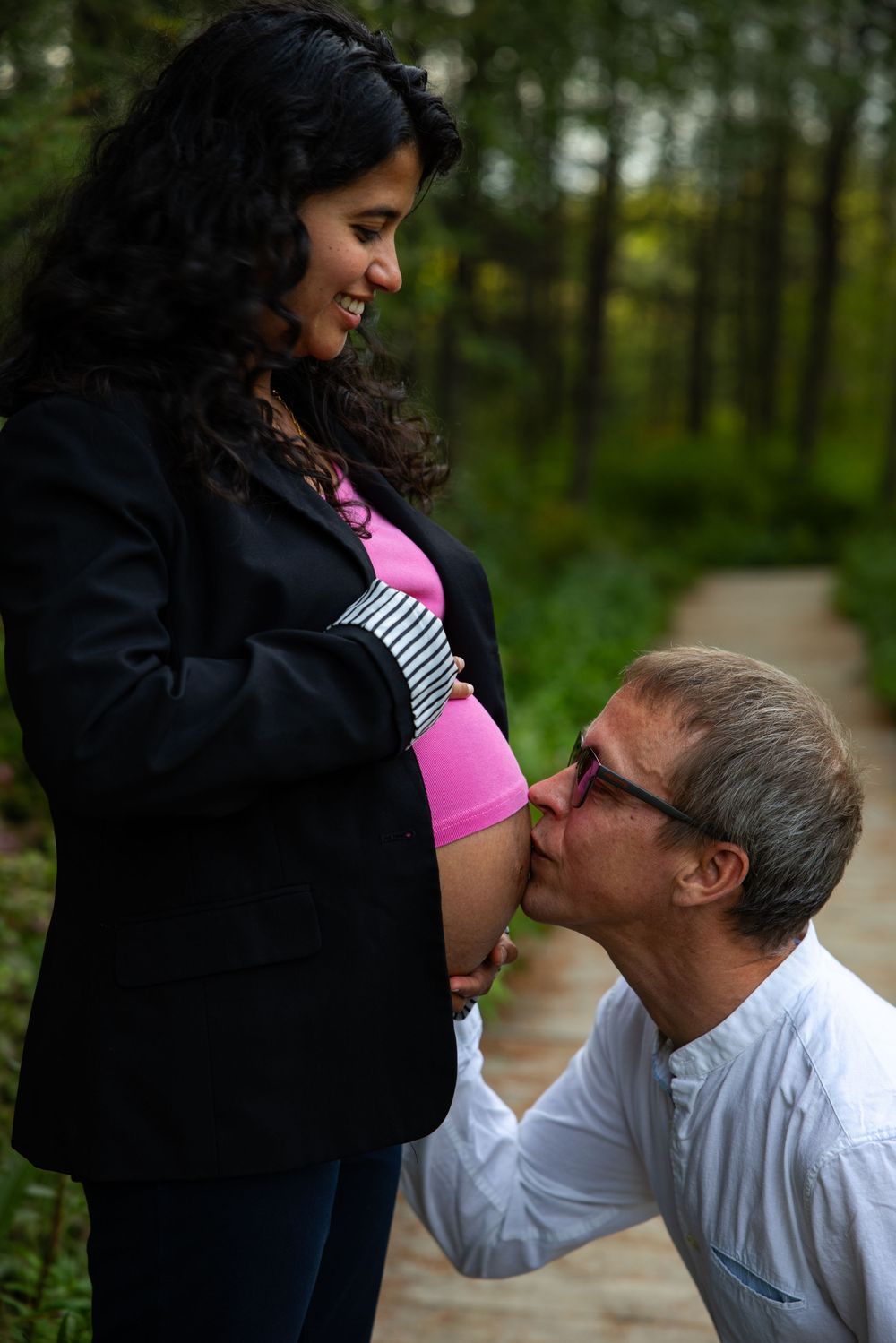 A tender maternity moment captured on a wooden path in the woods with a pink top and black jacket.
