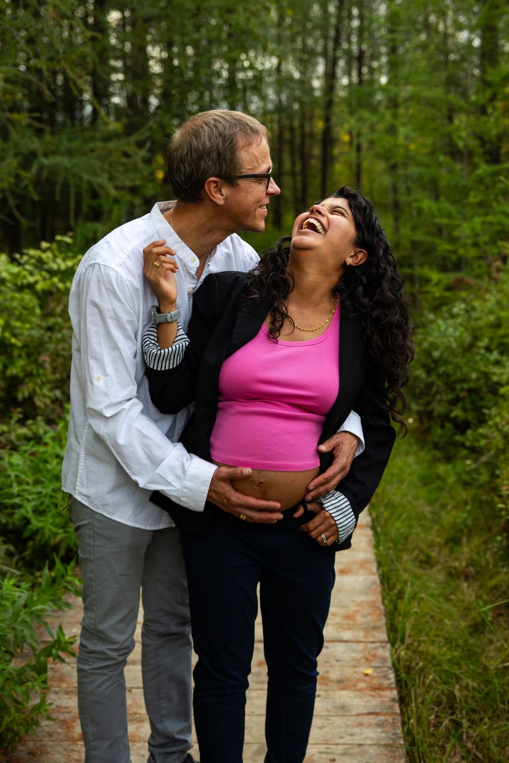 A couple shares intimate moments and laughter while posing together on a wooded path surrounded by greenery.