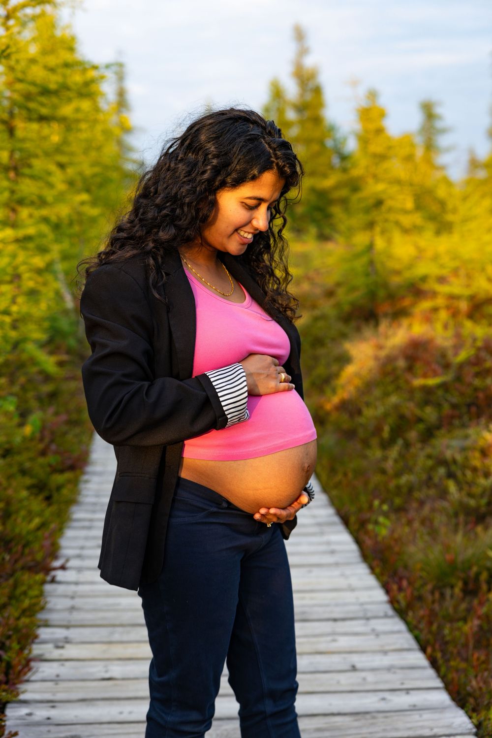 A pregnant woman in a black jacket and pink top stands on a wooden boardwalk surrounded by autumn foliage.