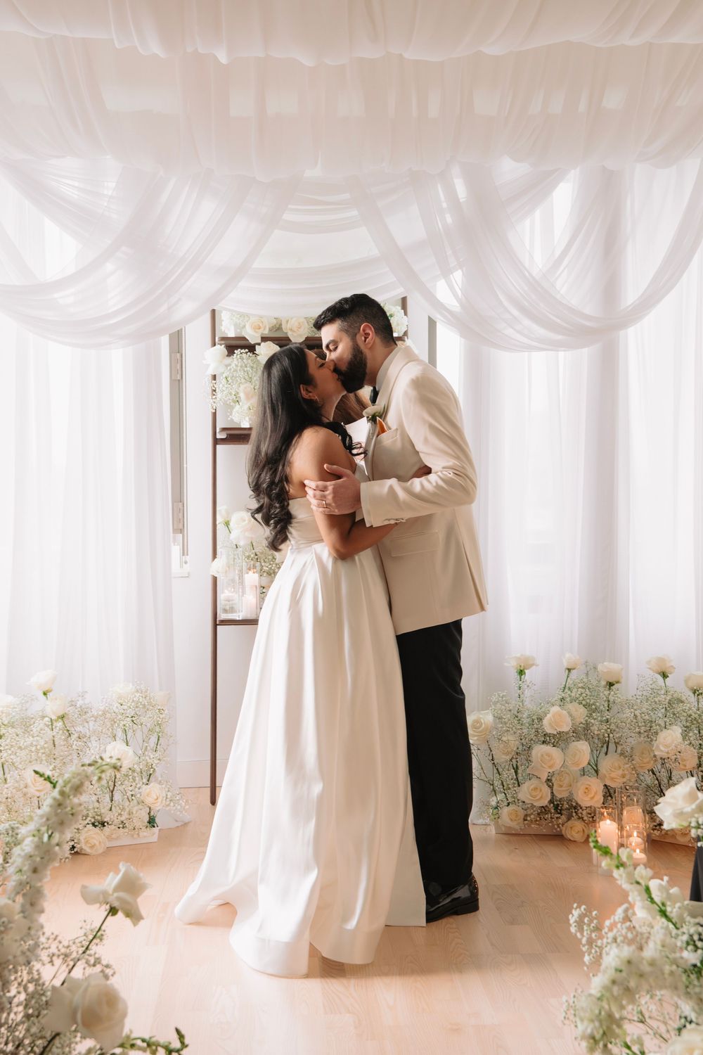 Romantic wedding moment captured in a light-filled room with white drapes and floral decor.