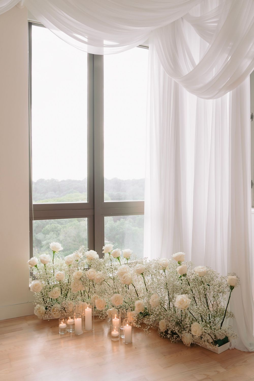 White floral arrangements and candles line windows with flowing white curtains.