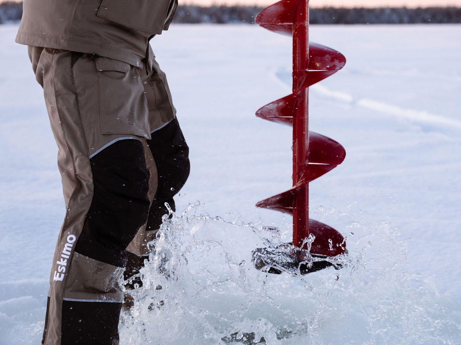 Red ice auger drilling through frozen lake surface with snow spray.
