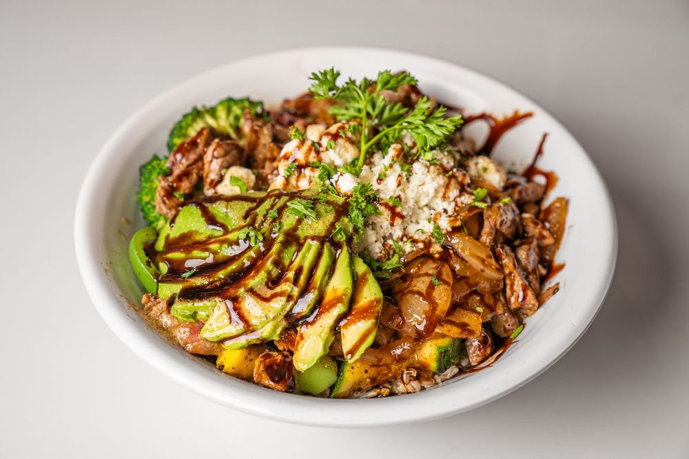 A white bowl filled with sliced avocado, broccoli, rice, seasoned meat, and drizzled with savory sauce.