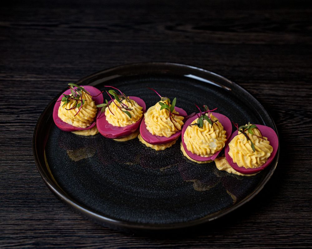 Five deviled eggs with pink-dyed whites and creamy yellow filling, garnished with microgreens on a dark ceramic plate.