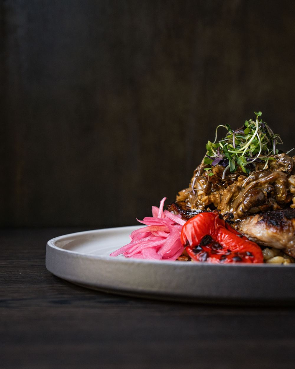 Gourmet roasted chicken dish with pickled red onions, roasted peppers, and fresh microgreens on a ceramic plate.