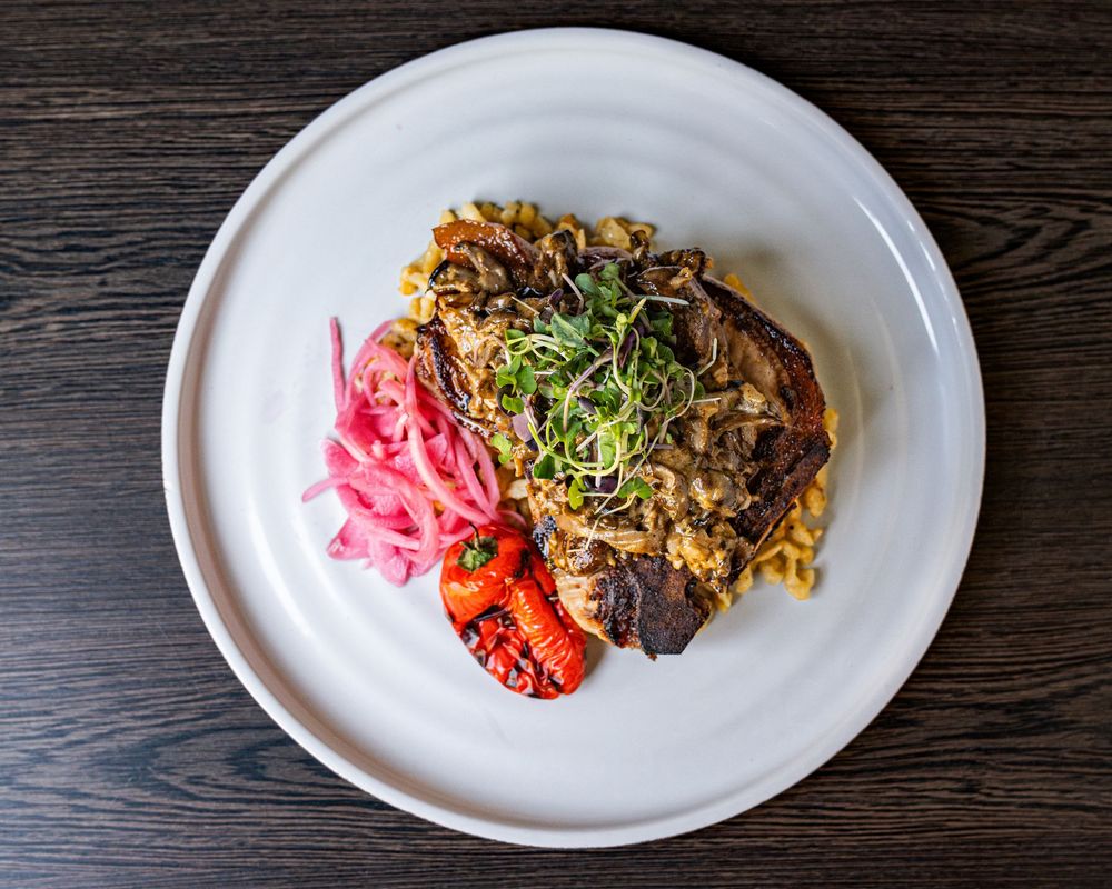 Overhead view of a gourmet meat dish with caramelized onions, microgreens, pickled red onions, and roasted peppers on a white plate.