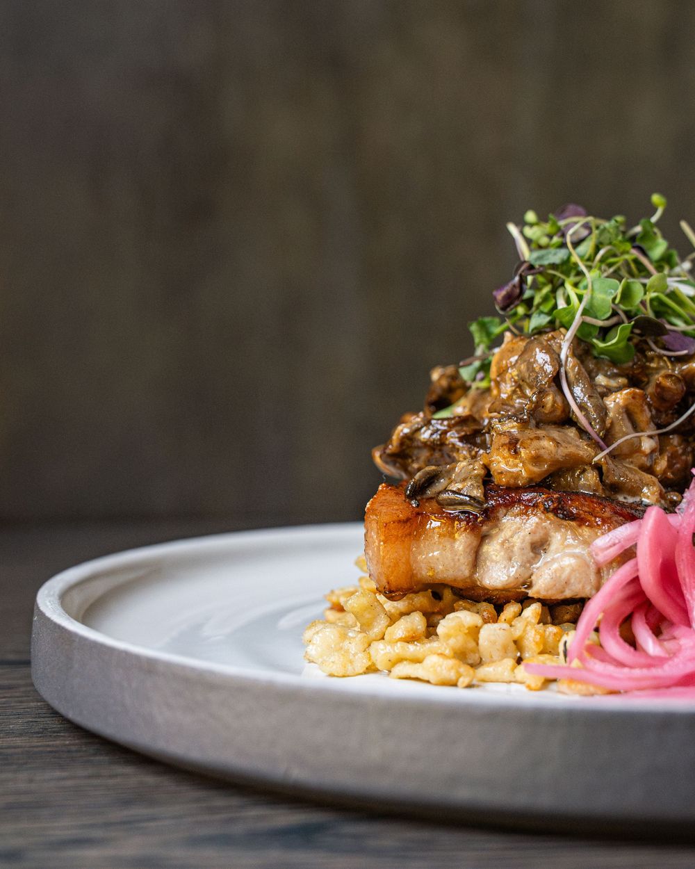 Gourmet pork chop served over creamy grits, topped with sautéed mushrooms, microgreens, and pickled red onions on a ceramic plate.