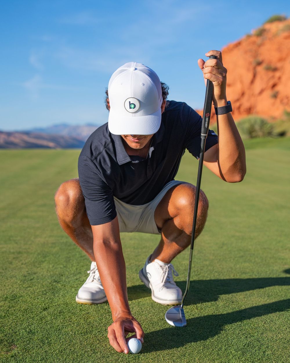 Golfer crouching on green course placing golf ball, wearing white cap, navy polo, and white shoes.