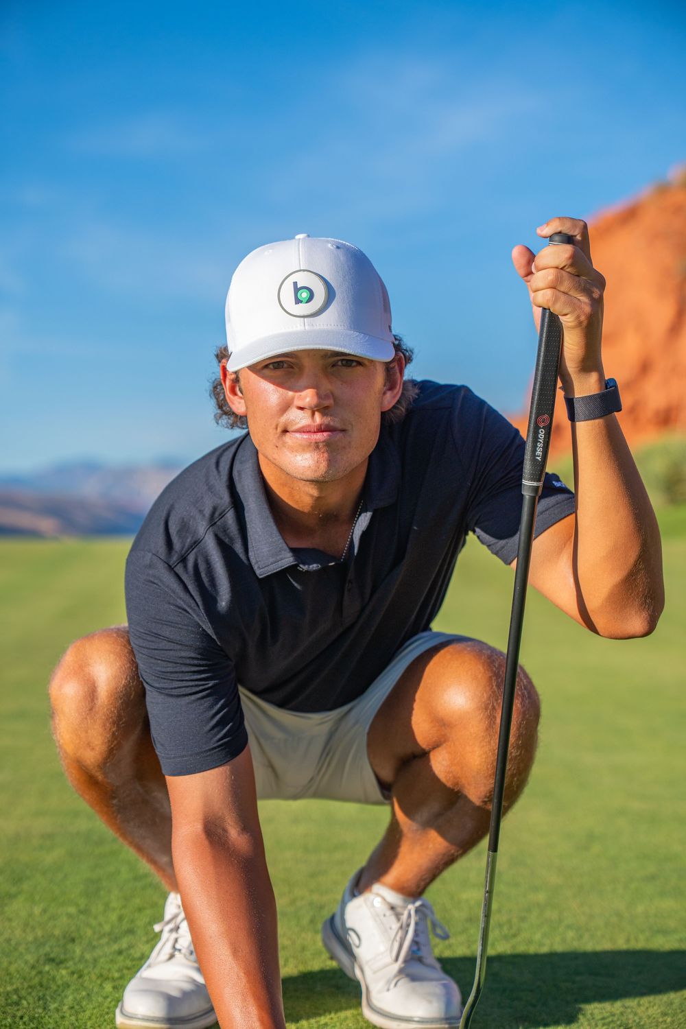 Young golfer crouching on green course, holding putter, wearing white cap and navy polo, with red rocks backdrop.