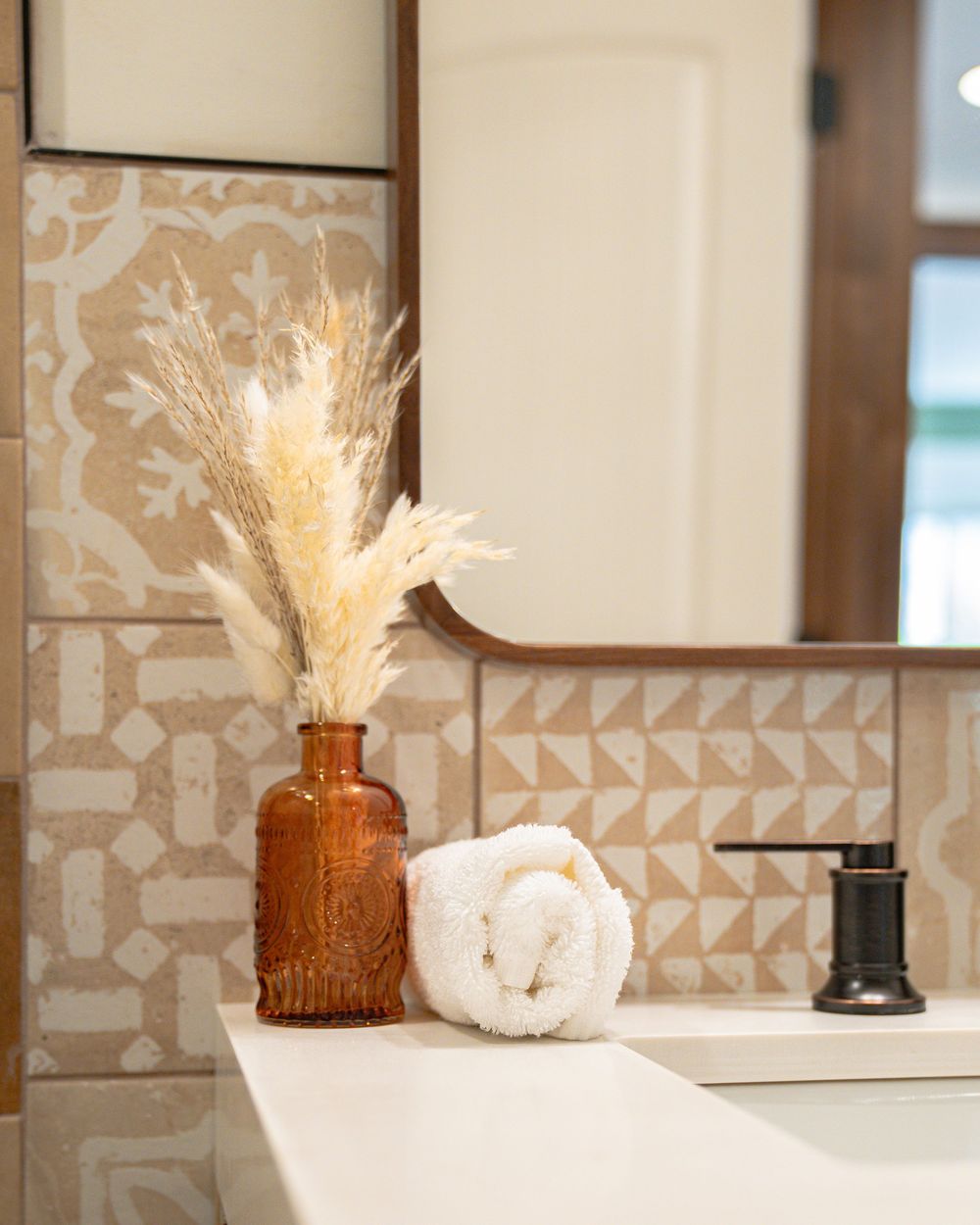 Decorative amber glass vase with pampas grass on white counter against geometric patterned wall tile.