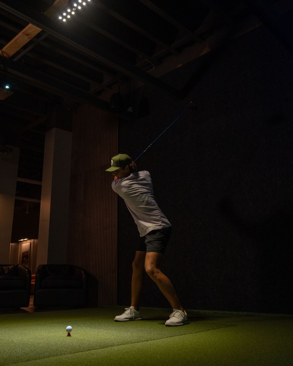 A golfer swings a club indoors at a dark golf simulator facility with green turf and bright overhead lighting.