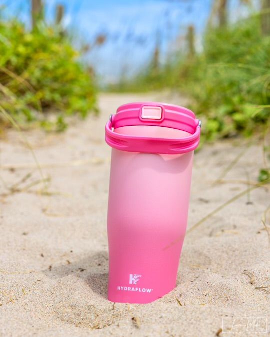 Pink reusable container positioned on sandy beach with green foliage in background.