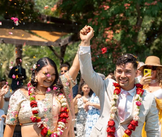 Wedding guests celebrate outdoors by throwing red rose petals during a joyful summer celebration.
