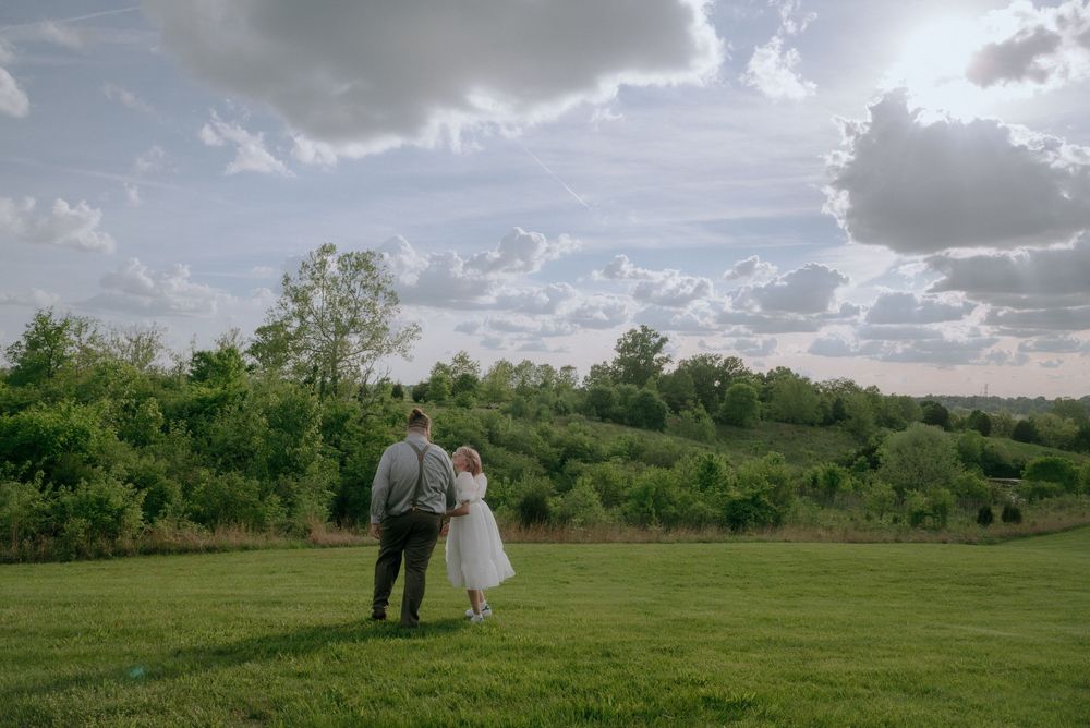 A romantic couple walks together across a scenic green field under dramatic clouds.