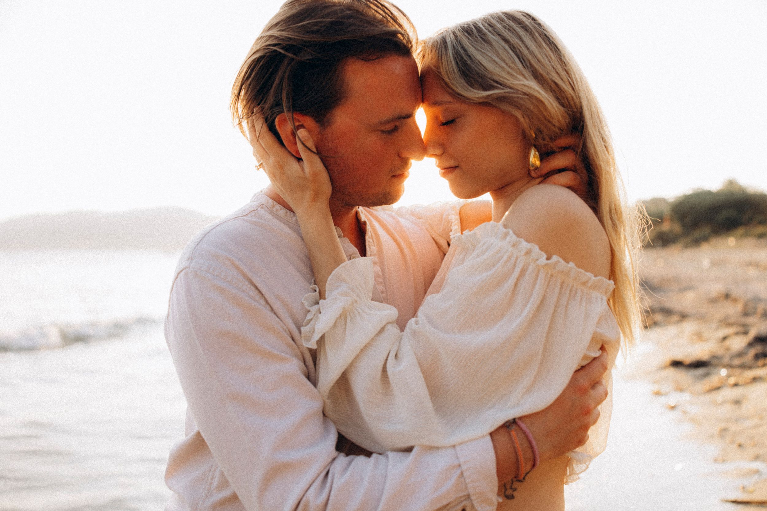 Romantic couple embraces on beach at sunset with ocean waves and coastline in background.
